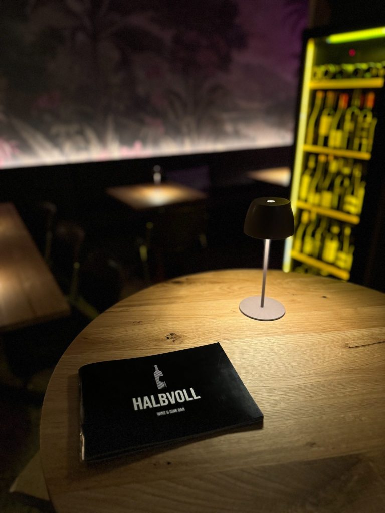 Tisch mit Holzoberfläche, einer Speisekarte von der Weinbar Halbvoll und einem Tischlicht, im Hintergrund ein Weinkühlschrank.