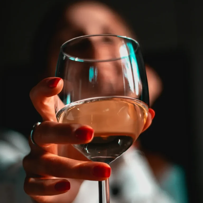 Ein Glas mit hellem Wein, gehalten von einer Person mit roten Nägeln im Vordergrund.