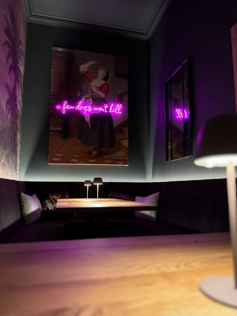 Hinterer Bereich in der Weinbar Halbvoll in Kassel mit einem Tisch, einer Lampe und einem neonbeleuchteten Kunstwerk an der Wand.