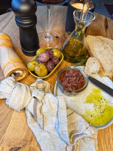 Ein gedeckter Tisch mit Oliven, Brot, Öl, Tomaten und einem Glas Wein.