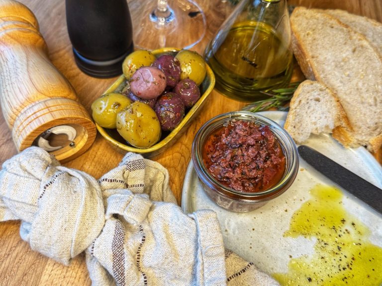 Ein rustikaler Tisch mit Brot, Olivenöl, Oliven, eingelegten Tomaten und Gewürzmühle.